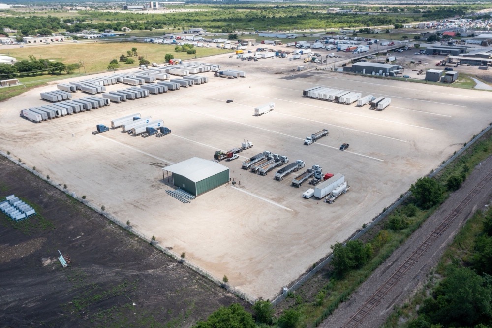 Aerial view of a large gravel truck and trailer parking yard with rows of parked trailers, tractors, and a small steel building.